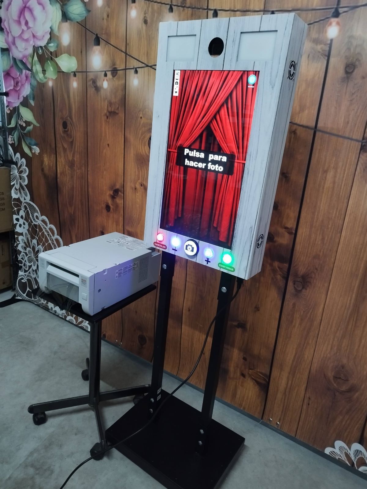 Fotomatón con pantalla táctil, impresora y fondo colocado contra una pared de madera y luces de colores.
