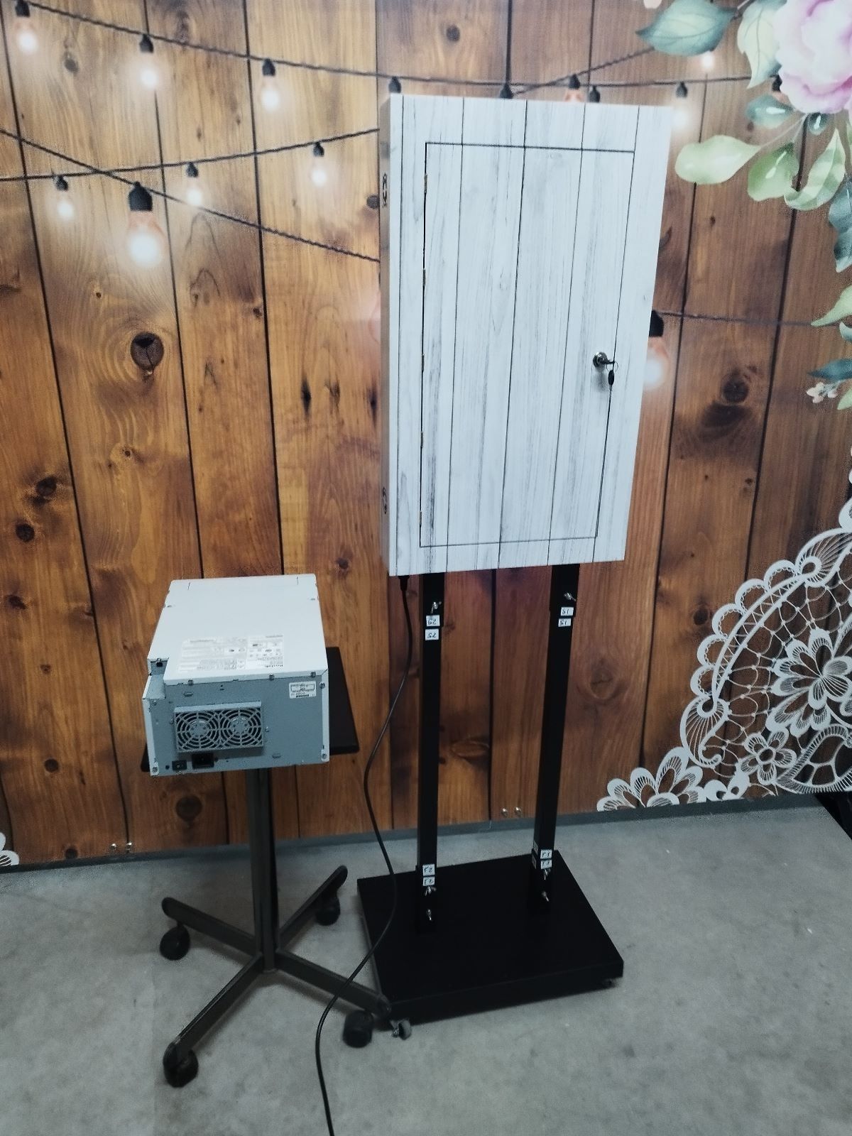 Configuración del fotomatón: gabinete estilo puerta de madera blanca sobre soporte negro, unidad de control sobre mesa rodante, fondo de madera.