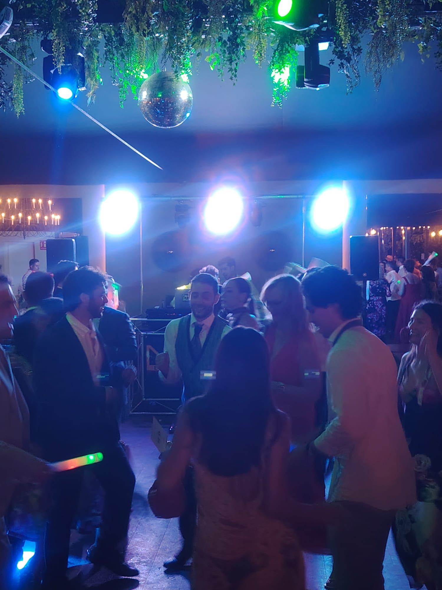 Gente bailando en una fiesta con luces, incluyendo una bola de discoteca. Luces verdes, azules y blancas iluminan la escena.