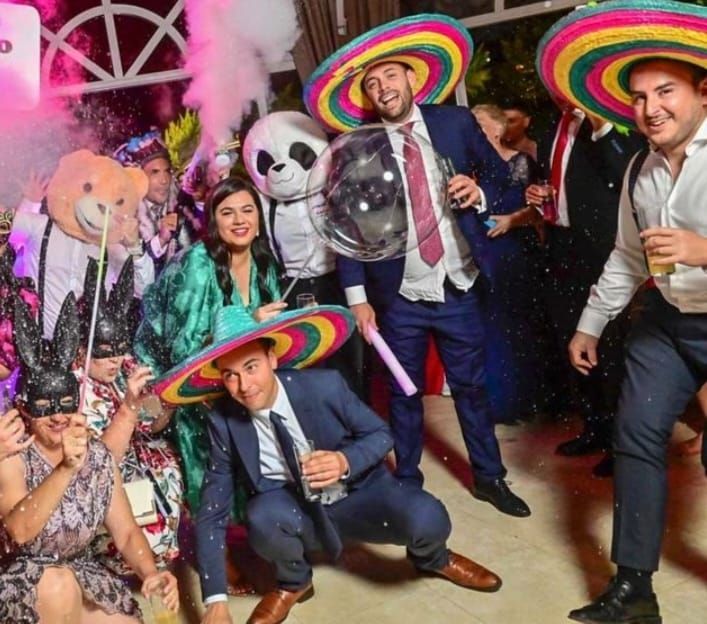Grupo de personas disfrazadas y con sombreros, celebrando en una fiesta, con confeti y humo en el aire.