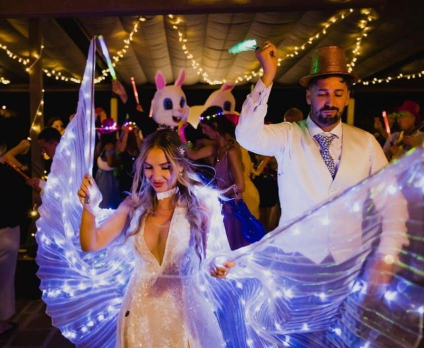 Los novios bailan en una fiesta con alas luminosas. Al fondo, gente con barras luminosas y una máscara de conejo.