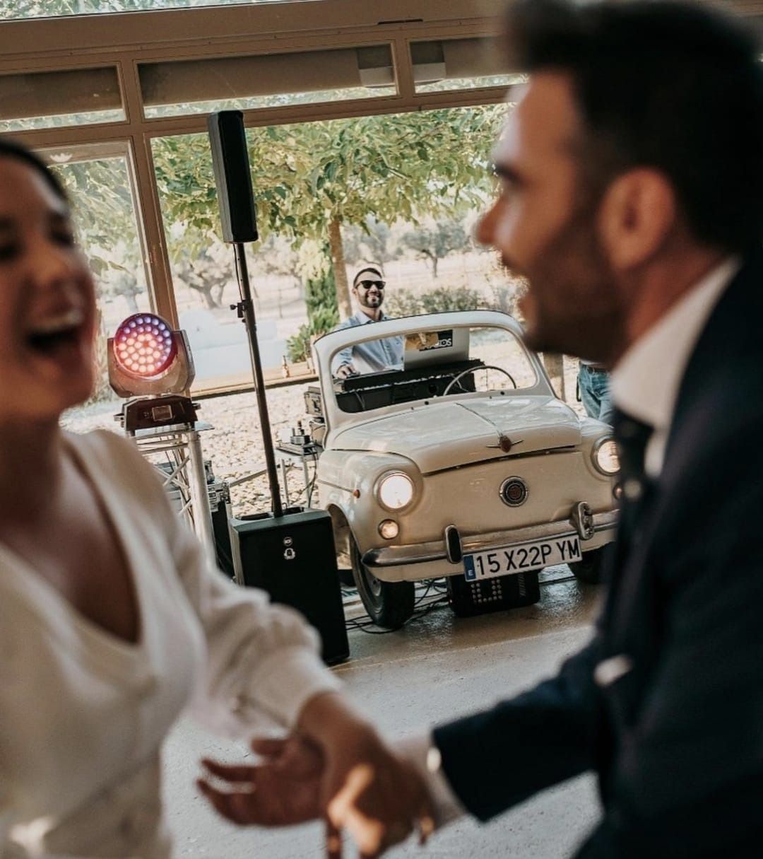 Los novios se ríen mientras bailan. DJ en un coche clásico, luces de fiesta y el lugar al fondo.