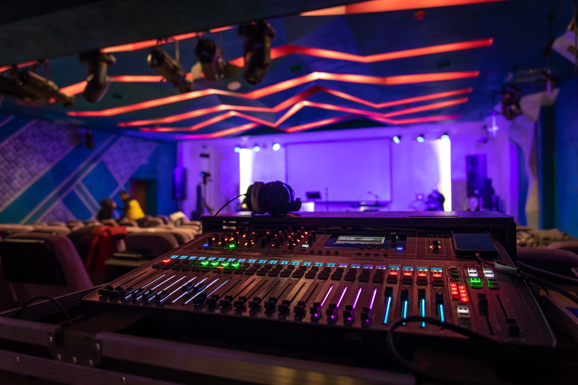 Consola de mezcla de sonido en un lugar poco iluminado con escenario; iluminación roja y azul.