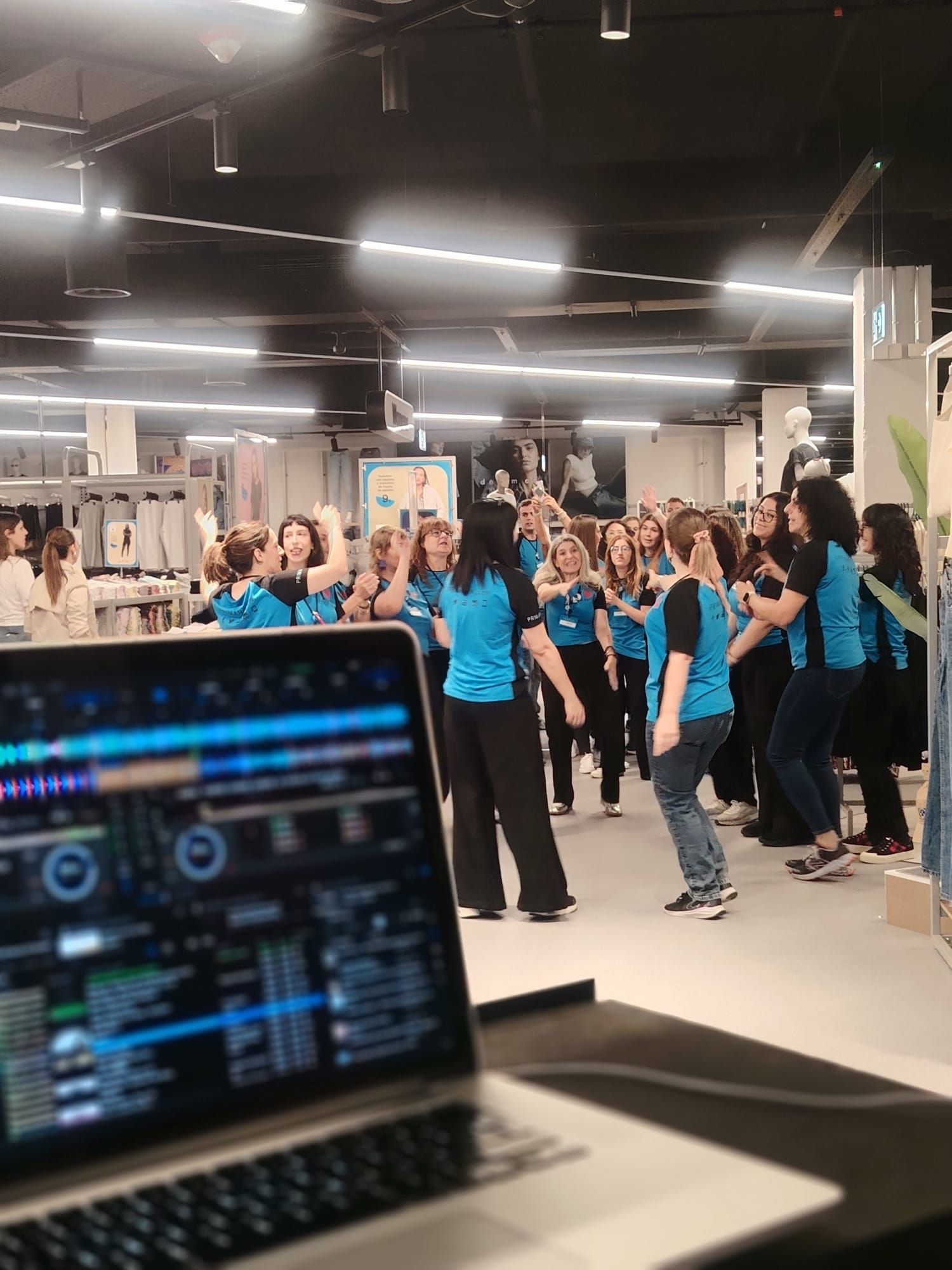 Una computadora portátil con software de DJ en primer plano; un grupo de personas bailando en una tienda, vistiendo camisetas azules.