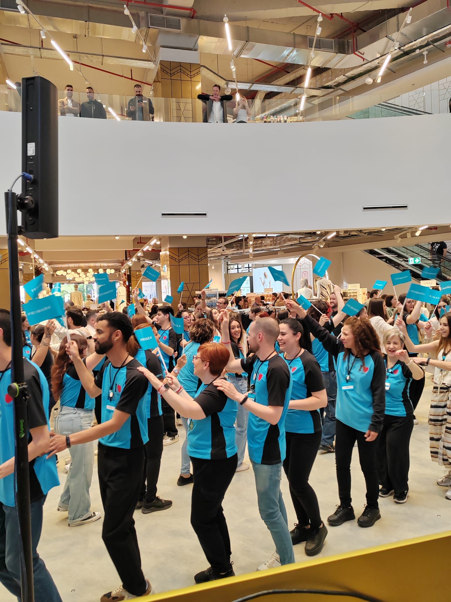 Personas con camisas azules bailando en una gran tienda con las manos levantadas, banderas y una audiencia en un balcón.