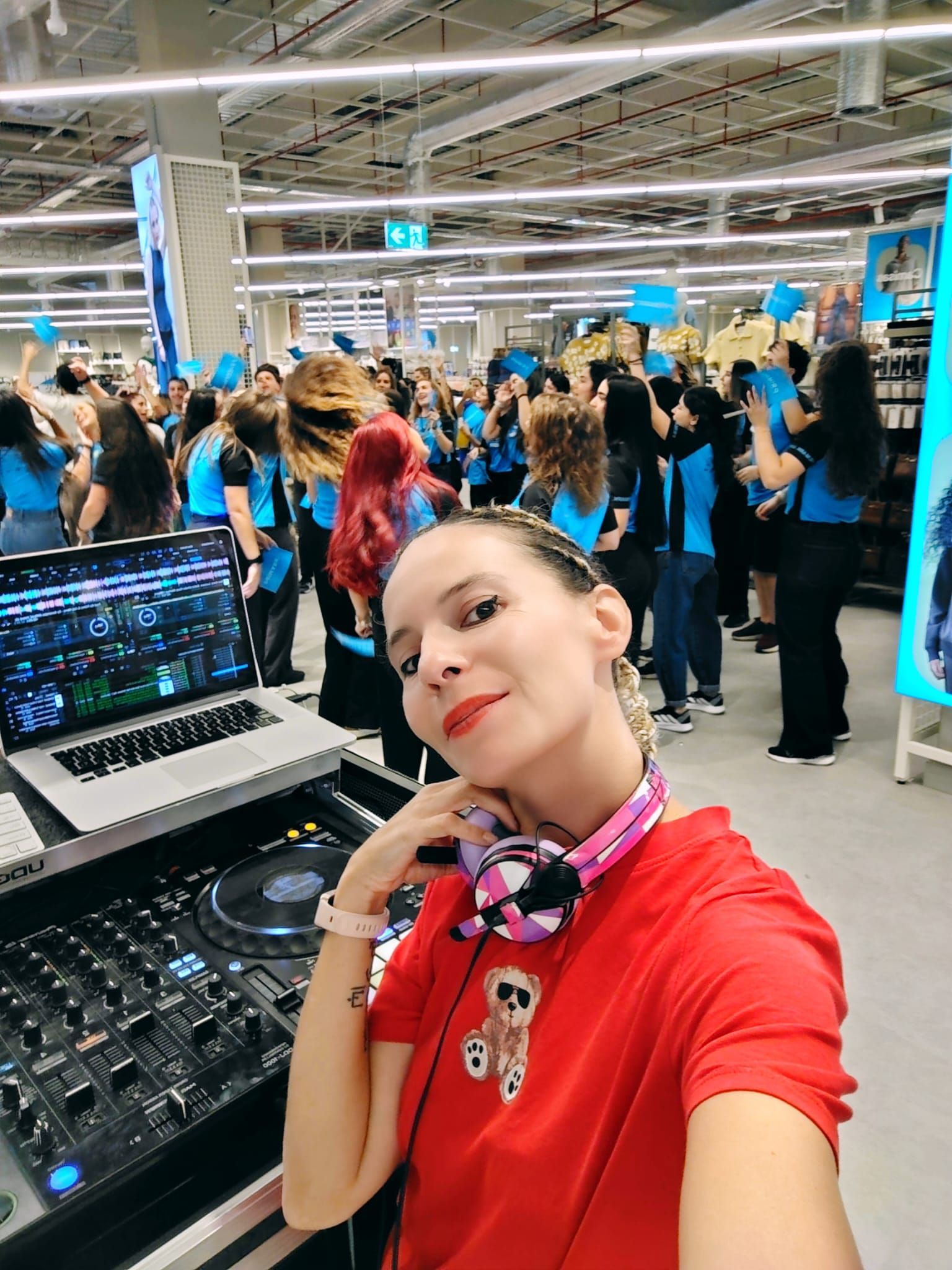 DJ en evento, sonriente, camiseta roja, auriculares, con multitud de personas en una tienda, detalles azules.