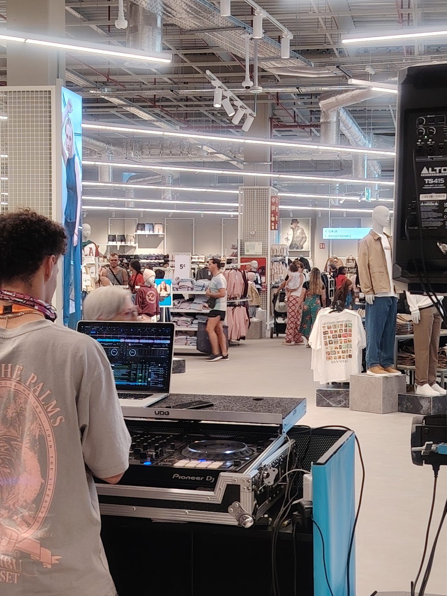 DJ en un evento en una tienda; tocando música con una laptop y otros equipos. Los compradores recorren los expositores de ropa.