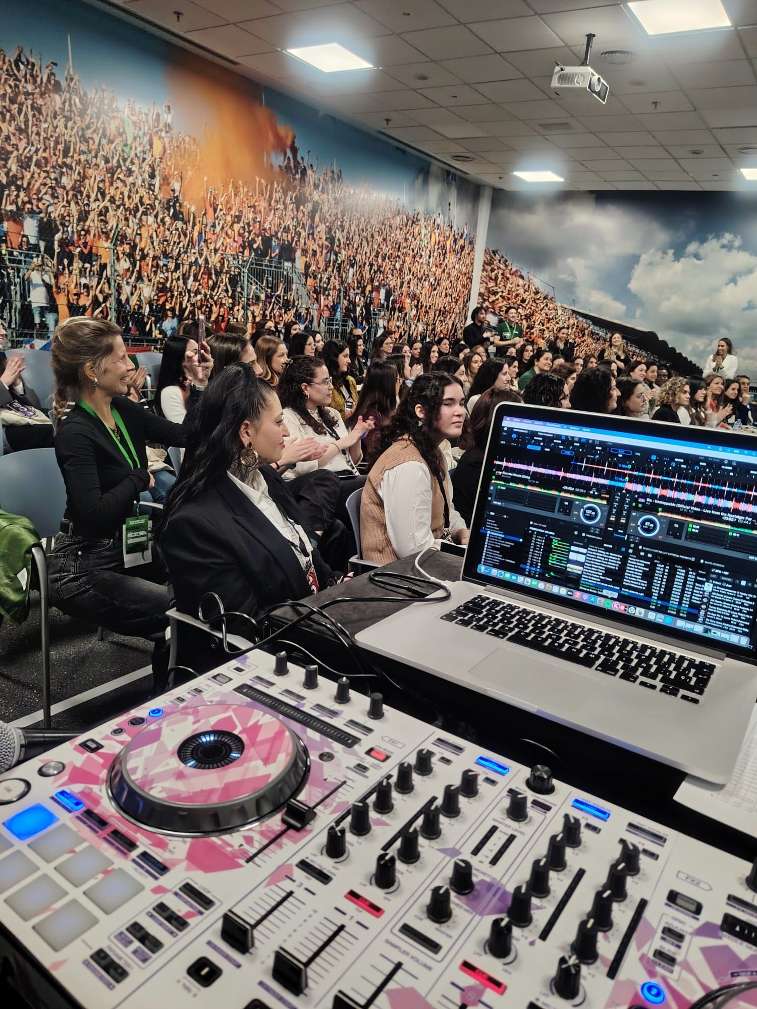 Configuración de DJ frente a un público sentado. Pizarra rosa y blanca, portátil con el software del mezclador, gente viendo un evento.