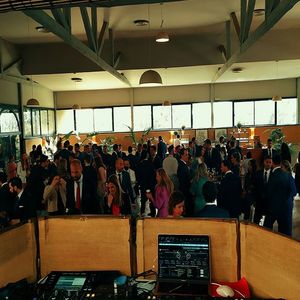 Recepción de boda con numerosos invitados en un gran salón. Cabina de DJ en primer plano, colores claros, grandes ventanales.