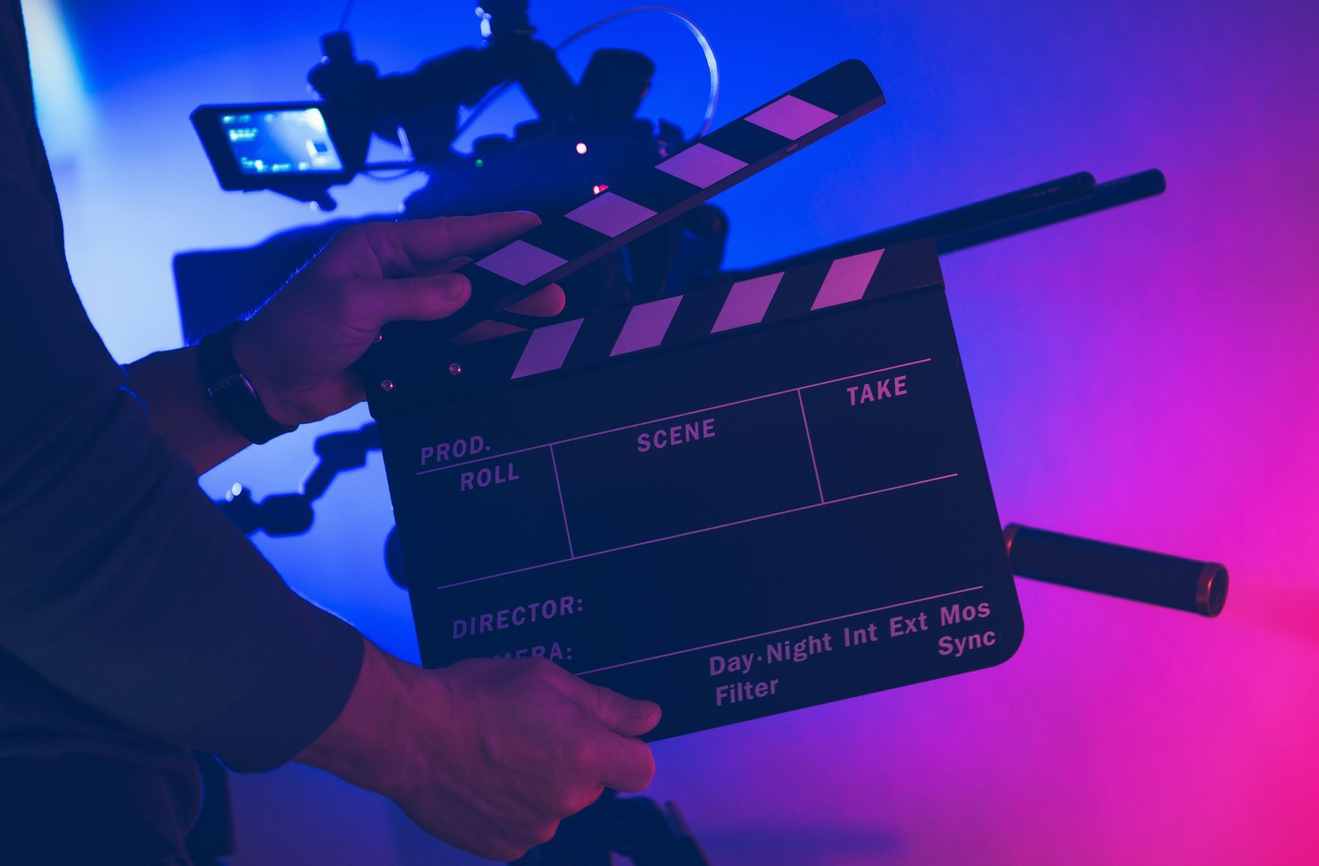 Persona sosteniendo una claqueta frente a una cámara de cine; iluminación azul y rosa.