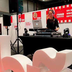 DJ en una feria de servicios de alimentación con laptop, mezclador, bocinas e iluminación de escenario. Una mujer sonríe con audífonos.