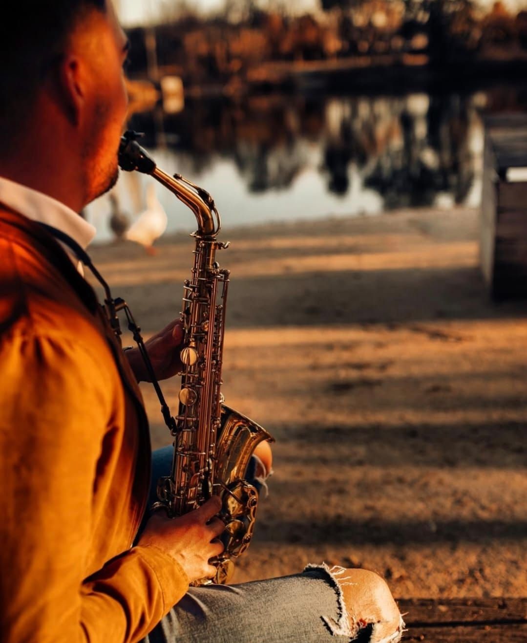 Un hombre toca el saxofón junto a un lago al atardecer. Una cálida luz dorada baña la escena.