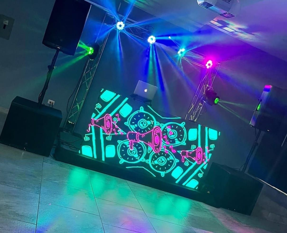 Configuración de DJ con altavoces, luces y una computadora portátil en una consola iluminada, con efectos de iluminación azul, verde y rosa.