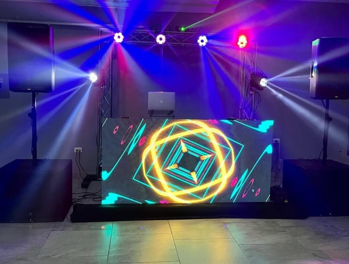 Configuración de DJ con pantalla LED, luces de escenario y altavoces. Rayos de luz azul y roja iluminan la sala.