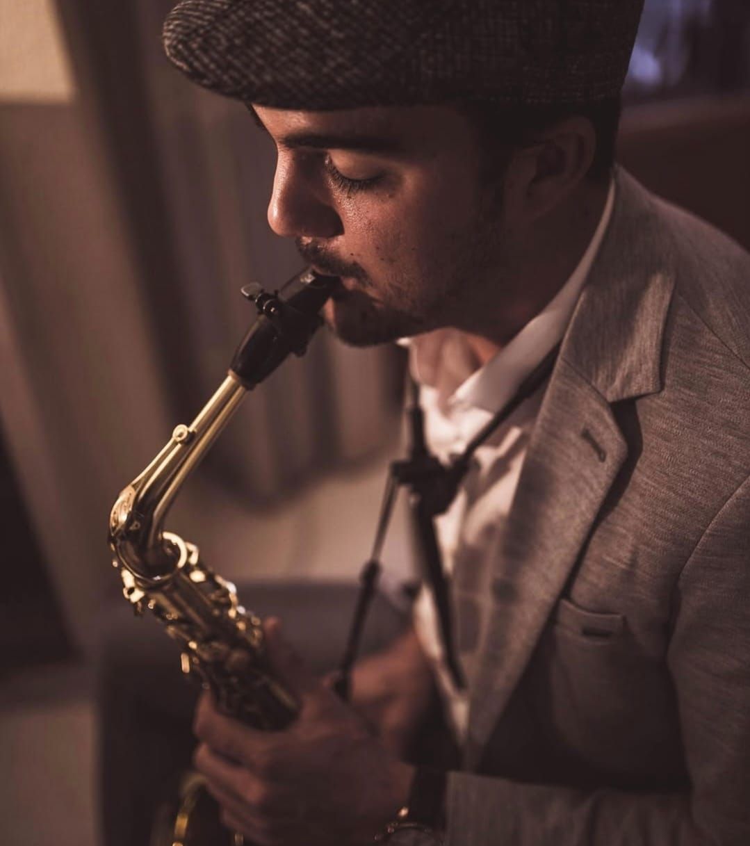 Hombre tocando un saxofón, vistiendo una gorra de tweed y una chaqueta, en un interior, expresión concentrada.