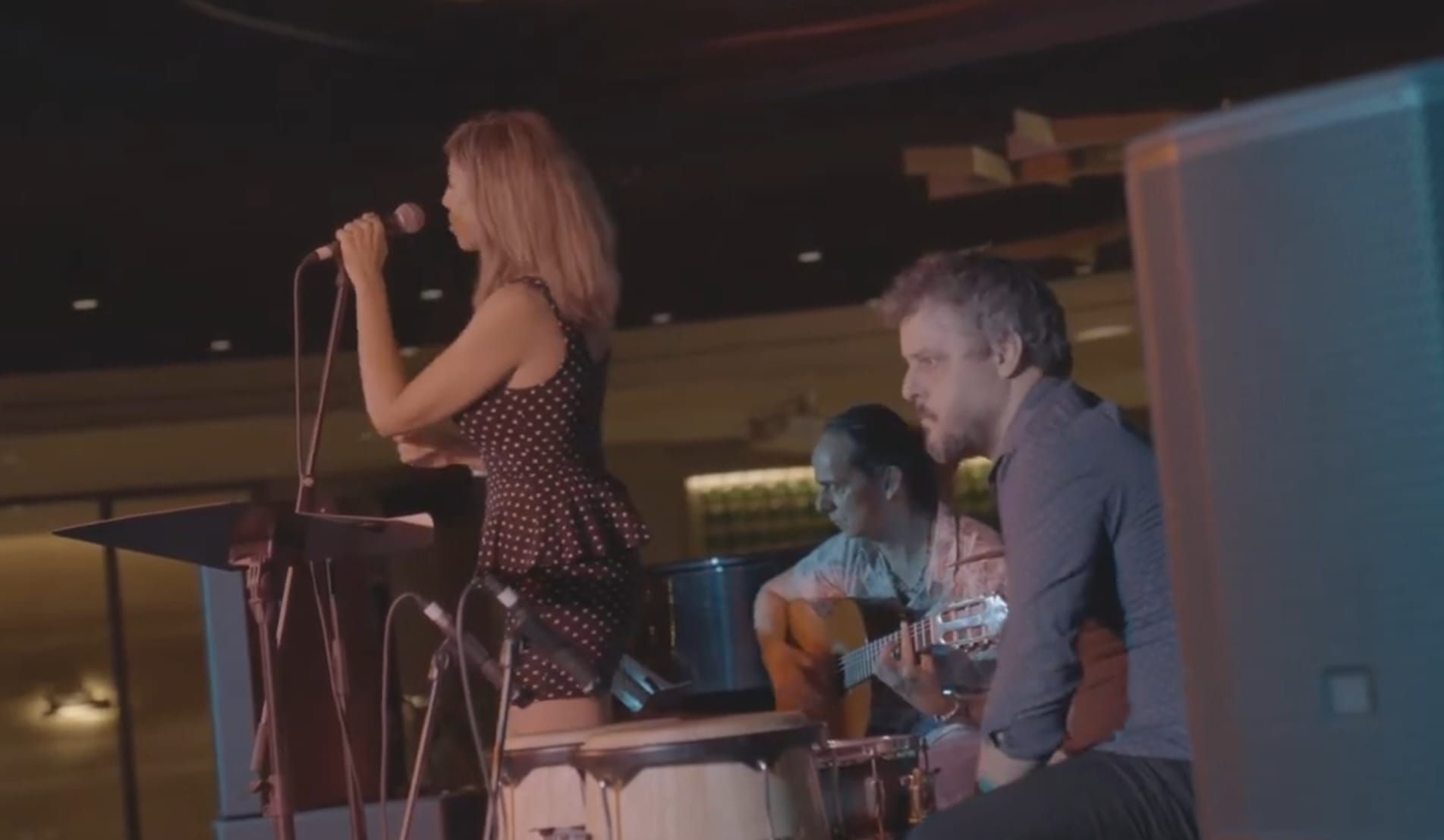 Mujer cantando en el escenario con una banda: percusionista y guitarrista. Iluminación tenue.