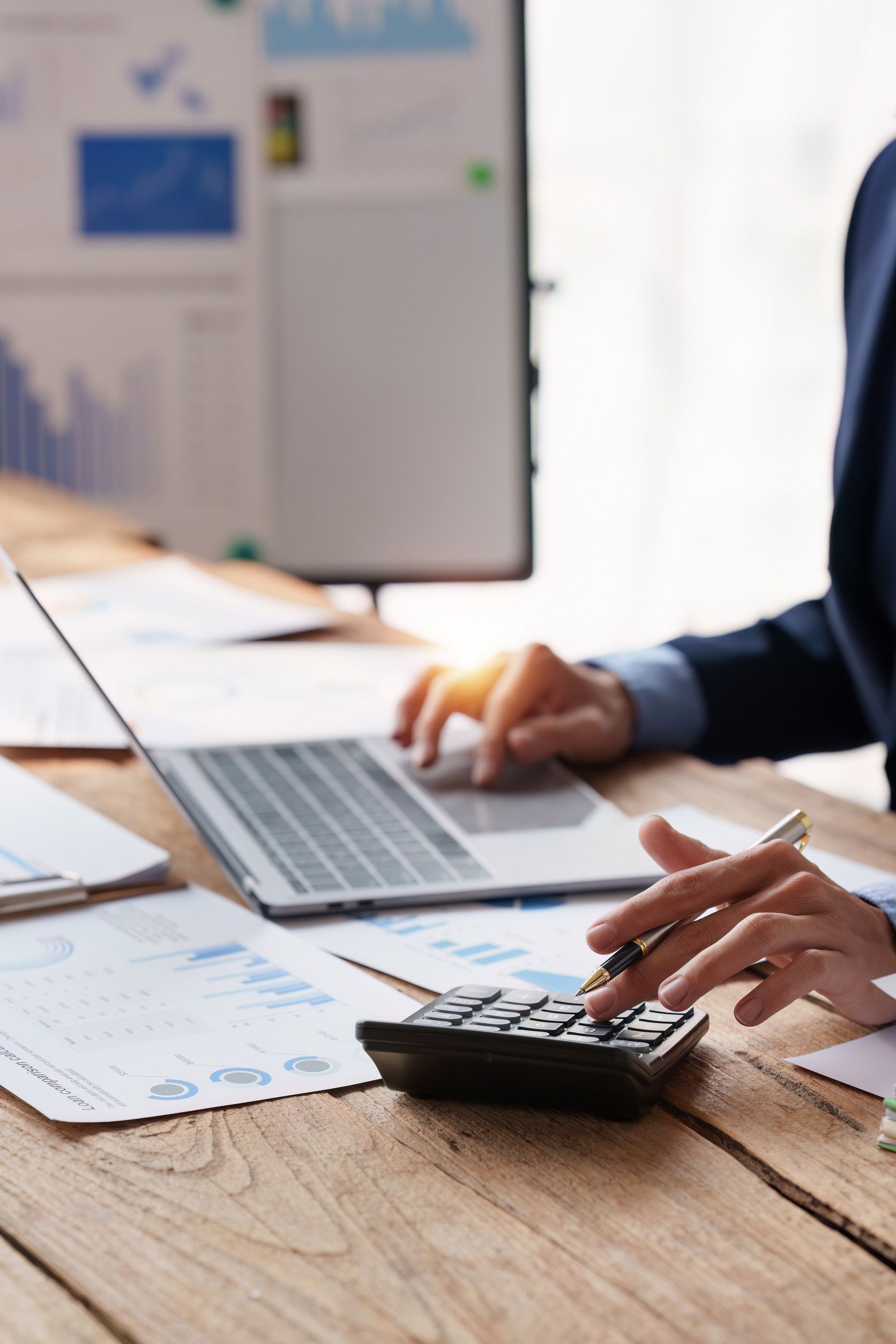 Persona con traje haciendo cálculos con calculadora en un escritorio con una computadora portátil y documentos financieros.
