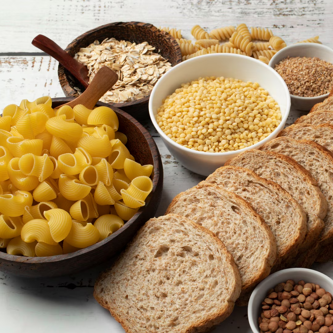 Cereali assortiti: pasta, pane, fiocchi d'avena, couscous, lenticchie e spirali su una superficie di legno.