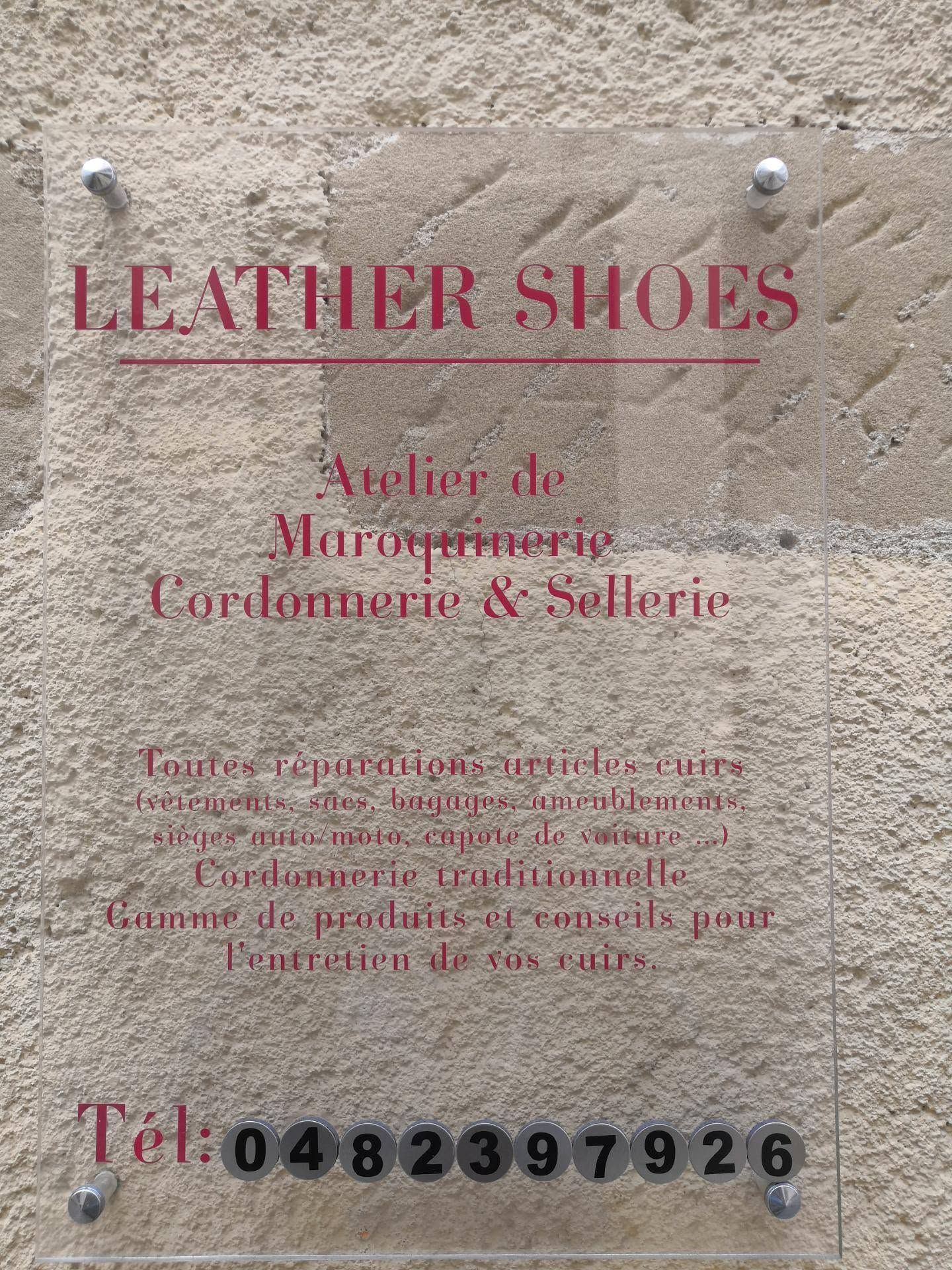 Enseigne de magasin de chaussures en cuir avec texte rouge sur un mur de pierre, listant les services proposés et un numéro de téléphone.