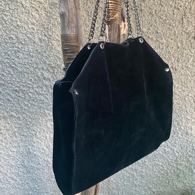 Sac à main en daim noir avec bandoulière chaîne, accroché contre un mur texturé.