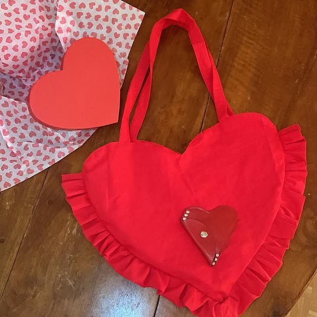 Un sac cadeau rouge en forme de cœur, aux bords froncés et orné d'un petit cœur, posé sur une table en bois.