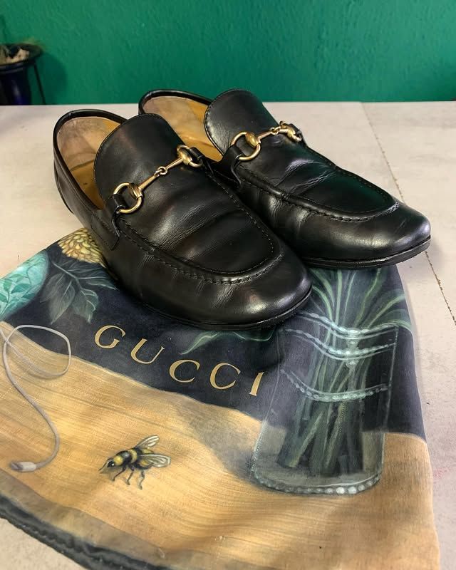 Des mocassins noirs ornés d'une chaîne dorée reposent sur une écharpe, elle-même posée sur un sol carrelé.