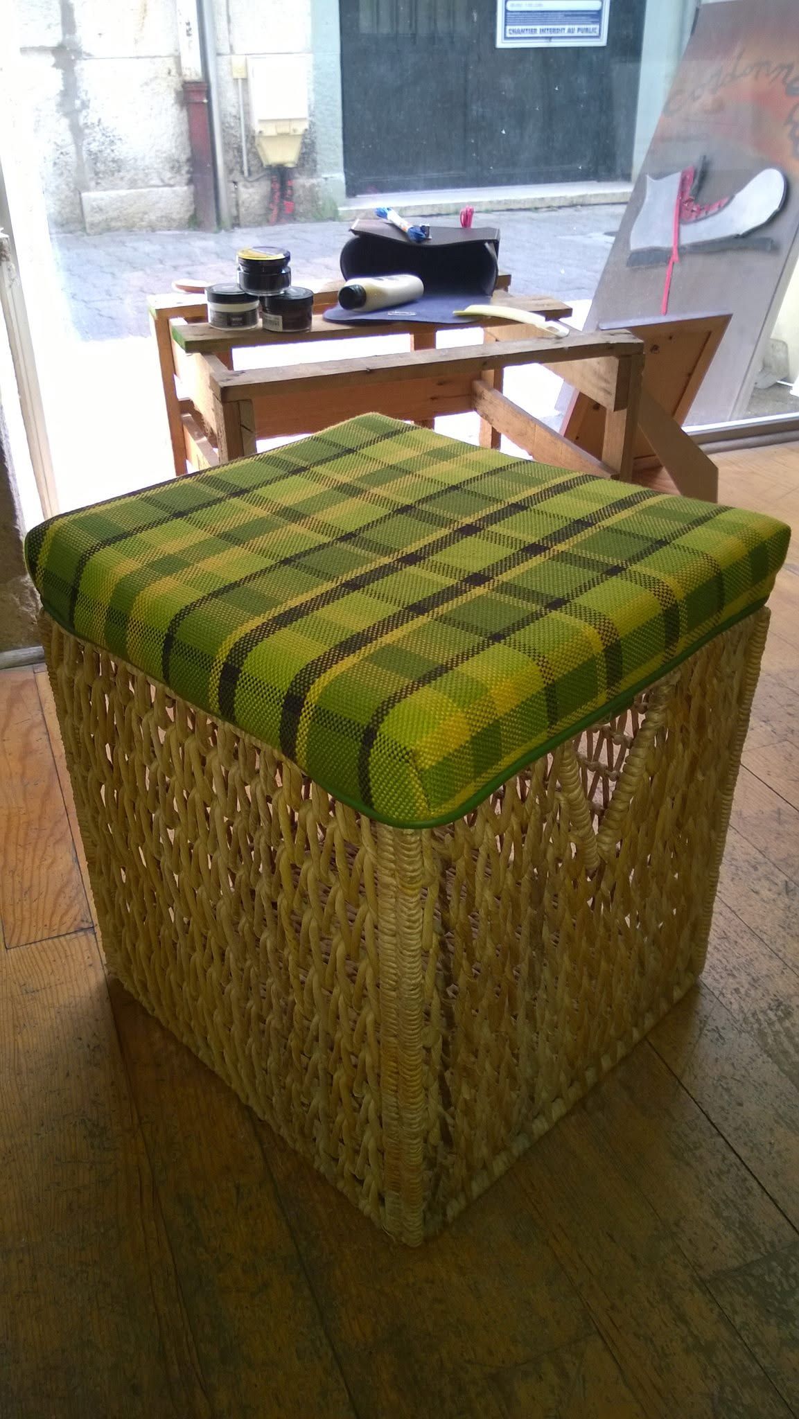 Un pouf à carreaux verts dans une pièce, avec une chaise en bois et une petite table près d'une fenêtre lumineuse.