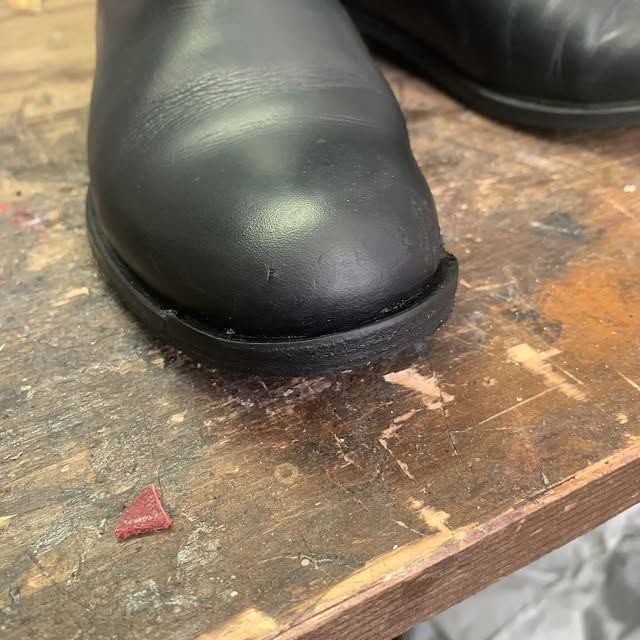 Bout d'une chaussure en cuir noir posé sur un établi en bois usé, avec un petit fragment rouge à proximité.