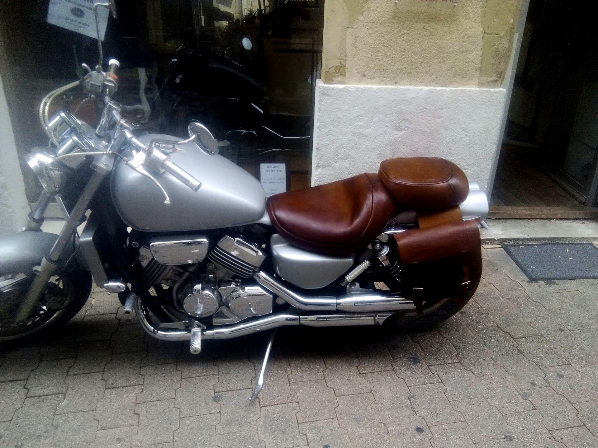 Moto argentée avec sièges en cuir marron garée dans la rue, à côté d'un bâtiment.