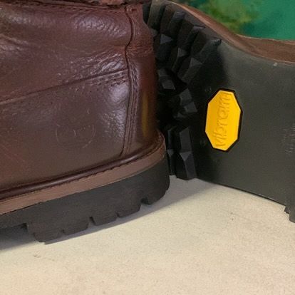 Botte en cuir marron à côté d'une semelle de chaussure de randonnée noire avec logo jaune sur un sol clair