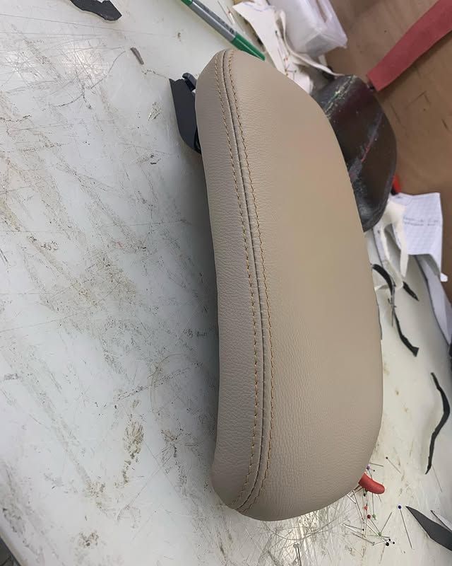 Assise de moto en cuir beige sur un établi, avec des outils de couture et des chutes de tissu à proximité.