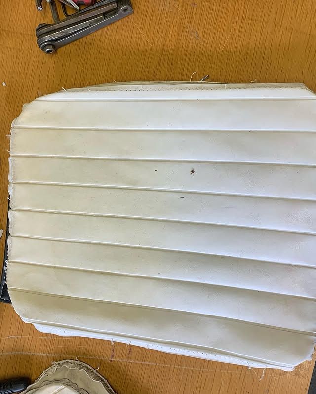 Coussin ou galette de siège blanc plissé sur une surface en bois, avec des outils partiellement visibles en haut à gauche.