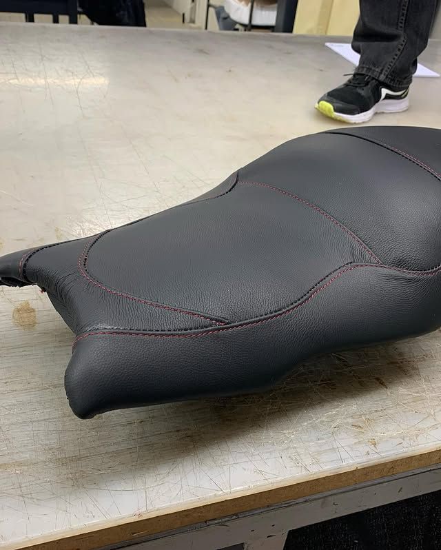 Selle de moto noire sur un établi, avec une personne debout à proximité, en arrière-plan.