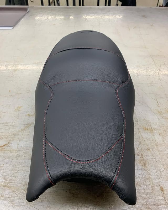 Selle de moto noire sur une table de travail, avec surpiqûres rouges et ouverture avant arrondie