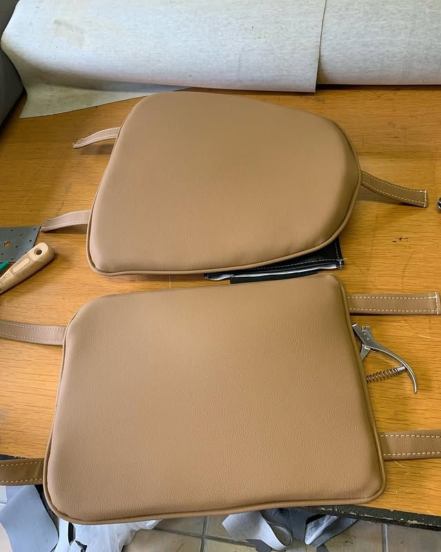 Deux coussins de siège beiges posés sur une table en bois, avec des sangles et des morceaux de mousse grise visibles à proximité.