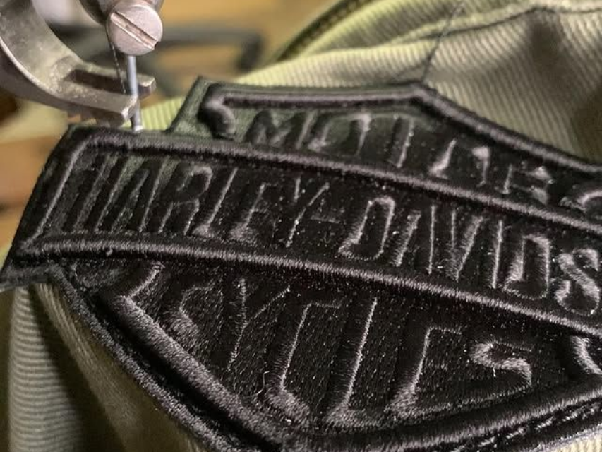 Gros plan sur un écusson Harley-Davidson noir sur un tissu olive, avec une fermeture éclair métallique à proximité.