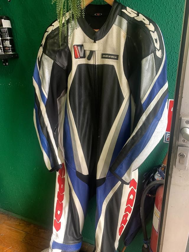 Combinaison de course moto noire, blanche et bleue accrochée à l'intérieur contre un mur vert.