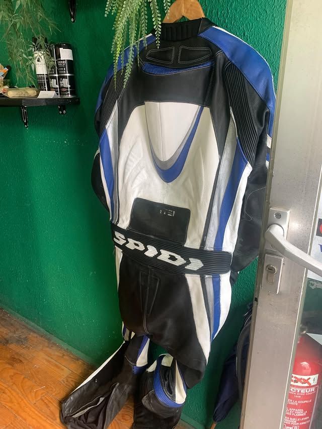 Combinaison de moto accrochée à un mur vert, avec des panneaux bleus, blancs et noirs.