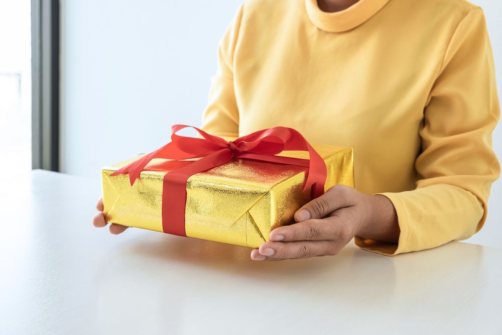Una mujer con una camisa amarilla sostiene una caja de regalo con una cinta roja.