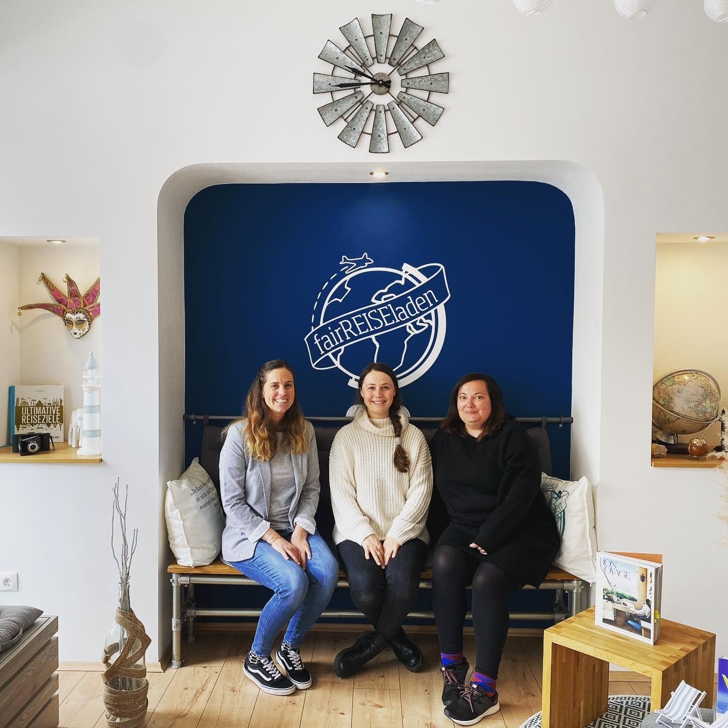 Drei Frauen sitzen auf einer Bank vor einer blauen Wand mit einer Uhr darauf.