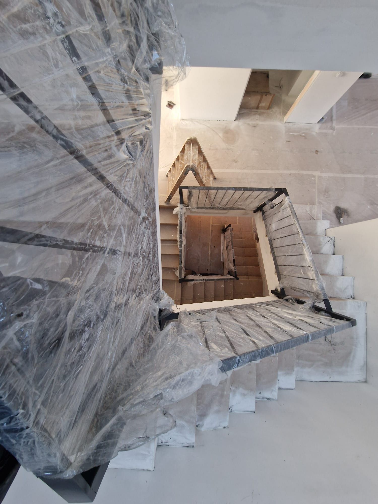 Escalera de caracol de madera vista desde arriba, envuelta en plástico protector en un interior blanco y luminoso.