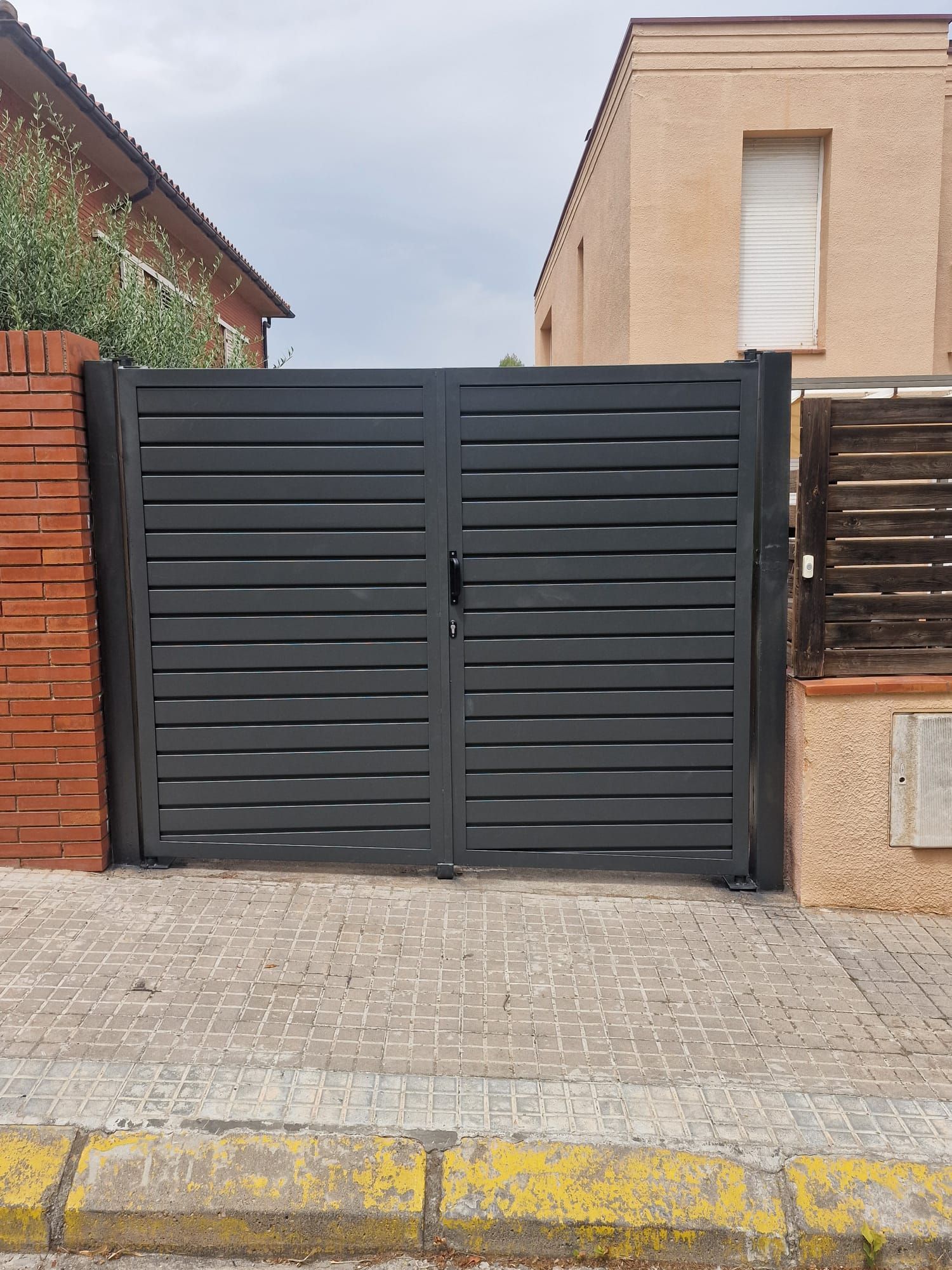 Portón negro de metal entre muros de ladrillo y casas beige en una calle residencial