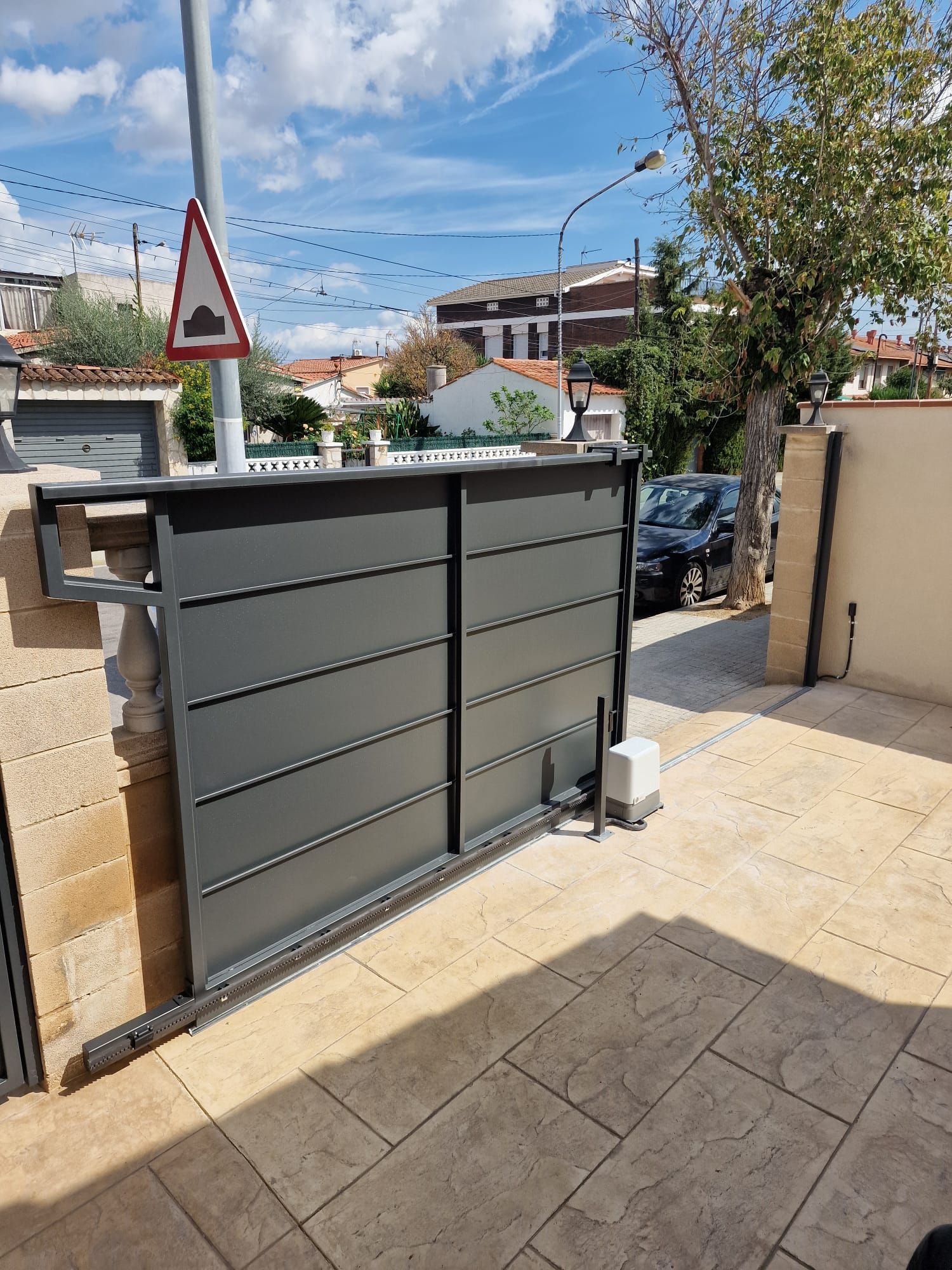 Portón gris de entrada a una casa, con una calle y coches aparcados al otro lado
