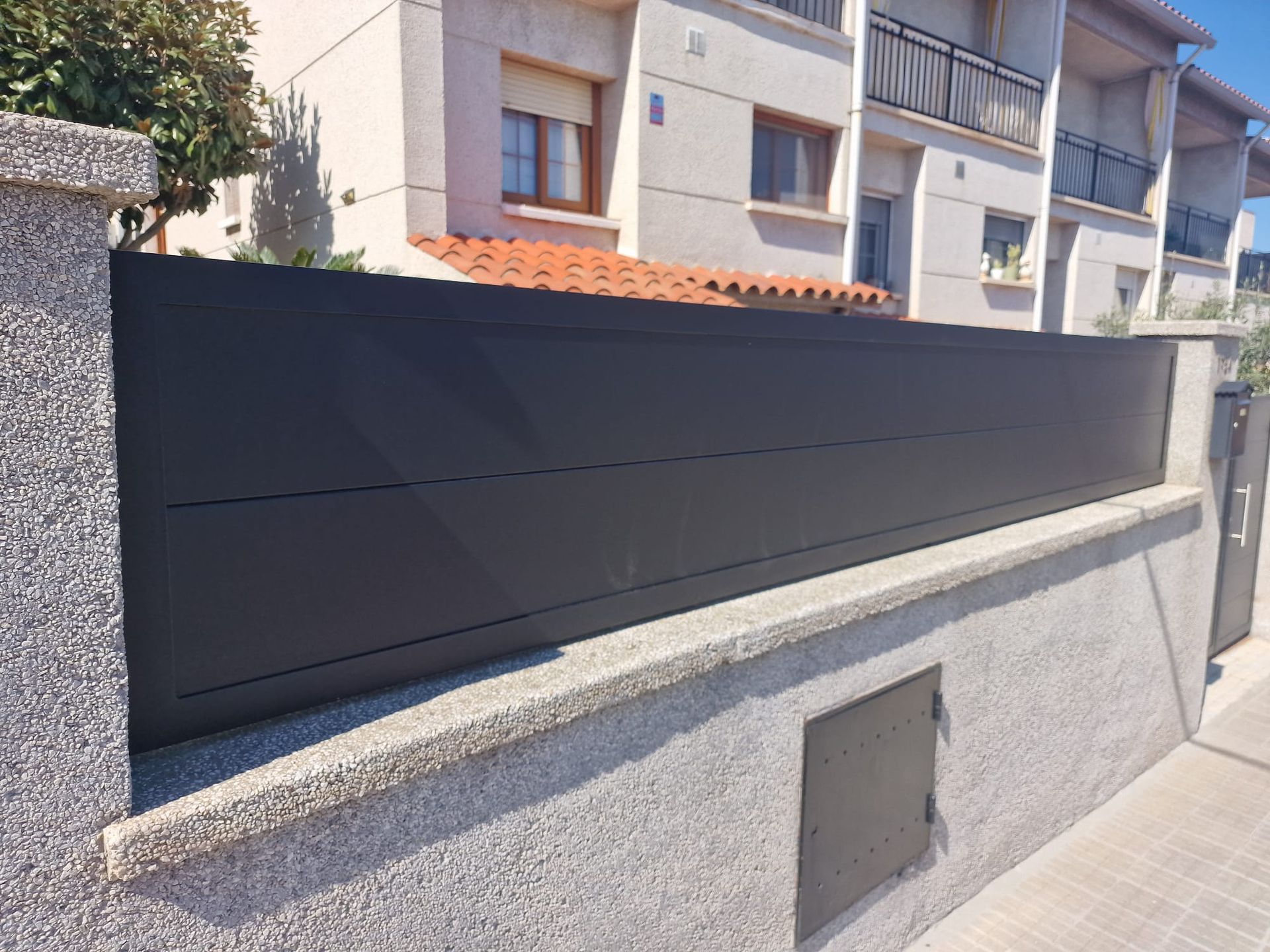 Valla negra de privacidad sobre un muro de piedra gris junto a una acera residencial