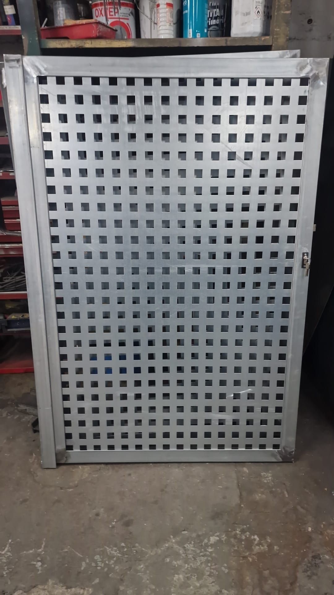 Panel metálico gris perforado apoyado contra unas estanterías en un taller o zona de almacenamiento.
