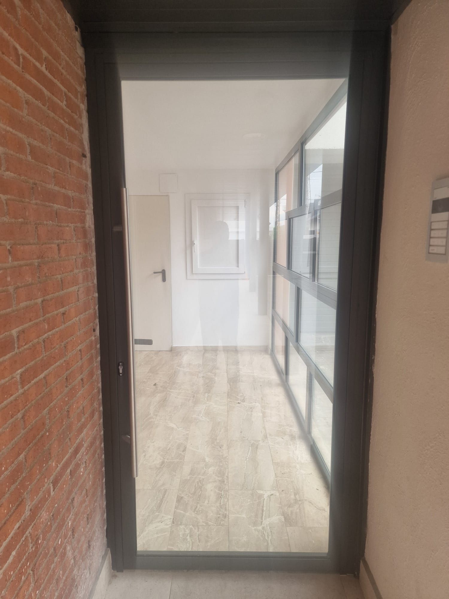 Pasillo estrecho con una pared de ladrillo, una puerta de cristal y suelo de madera clara que da a un interior luminoso.