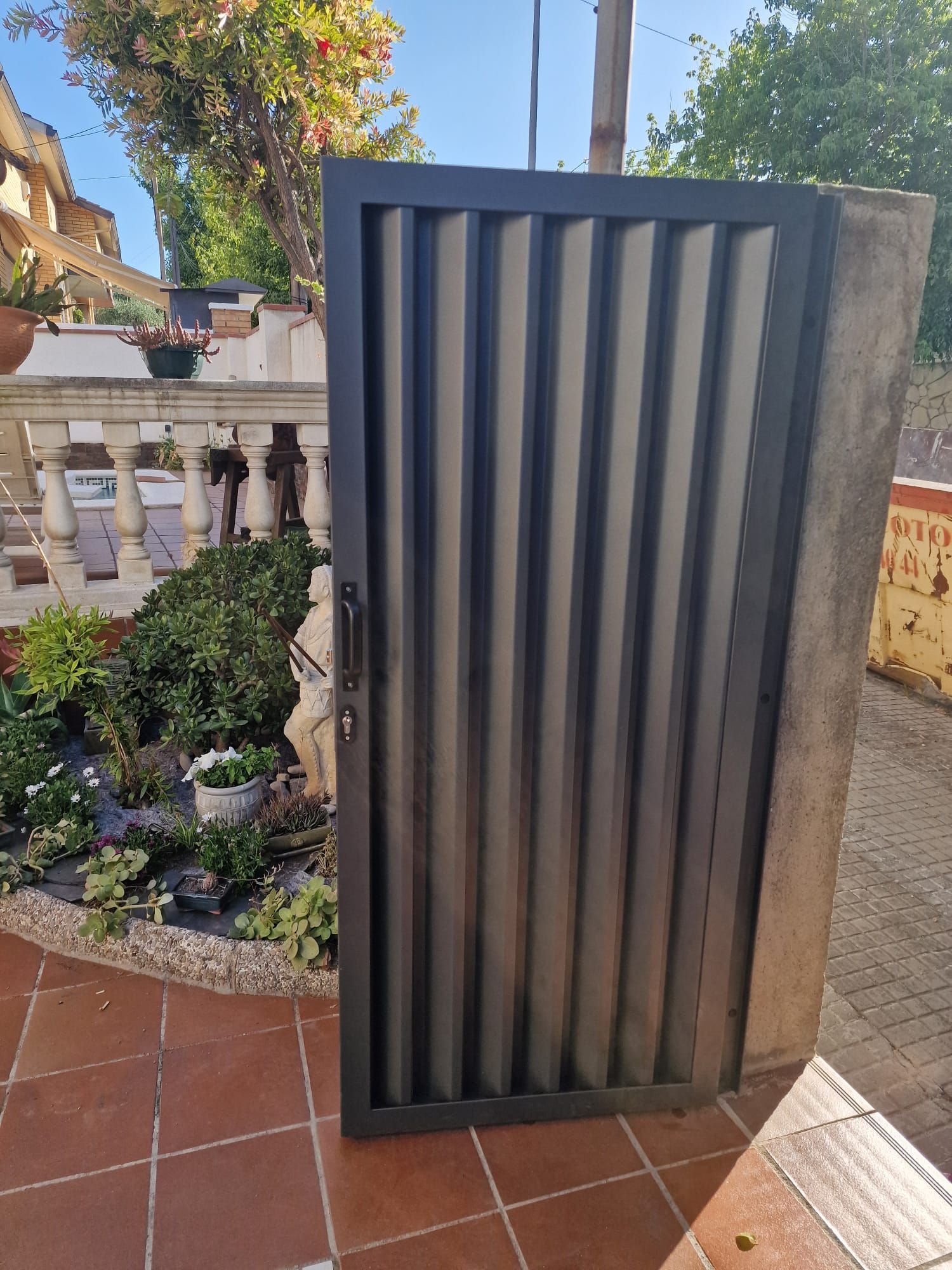 Pantalla de privacidad negra de listones en un patio embaldosado, junto a un jardín y una barandilla de piedra