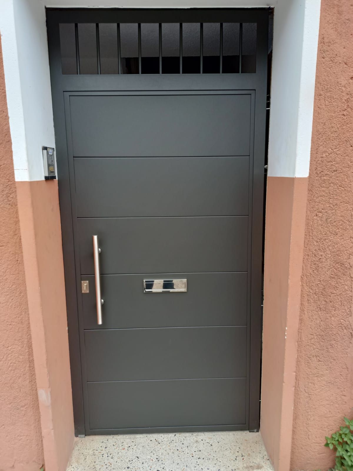 Puerta principal gris oscuro con un tirador vertical y una ranura para el correo, en una entrada en tonos melocotón y blanco