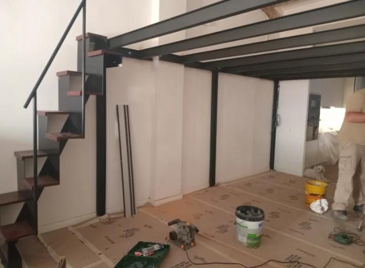 Interior en construcción con una escalera de metal negro, vigas de altillo y herramientas sobre un suelo.