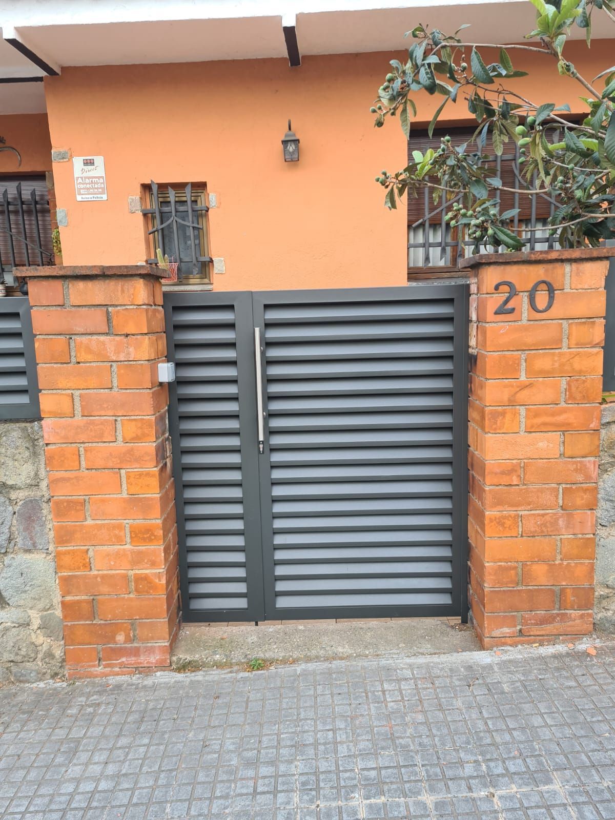 Entrada de una casa naranja con una verja metálica gris entre pilares de ladrillo y una acera empedrada